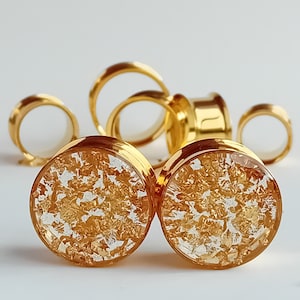 May include: Gold-colored ear gauges with a clear center filled with gold leaf flakes. Several gold-colored tunnels and rings are in the background. The gauges have a shiny, metallic finish and a circular shape.
