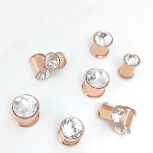 May include: Rose gold-toned ear gauges, each featuring a clear, faceted crystal. The jewellery is arranged on a white background. The gauges vary in size and position, highlighting their design and sparkle.