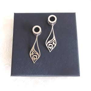 Puede incluir: Un par de túneles para oreja de metal plateado con colgantes en forma de lágrima. Cada colgante tiene un intrincado patrón de filigrana calada con líneas curvas. Los túneles son circulares con un centro hueco para dilataciones.