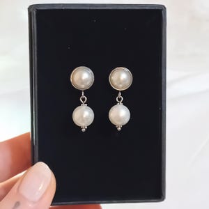 Può includere: Un paio di orecchini pendenti con perle. Ogni orecchino presenta un piccolo perno di perla rotondo con una perla pendente più grande. Gli orecchini sono esposti in una scatola nera.