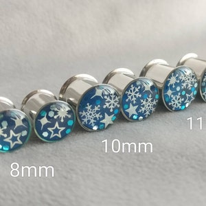 May include: Set of six silver ear gauges with blue and white snowflake designs. The gauges are 8mm, 10mm, and 11mm in size.