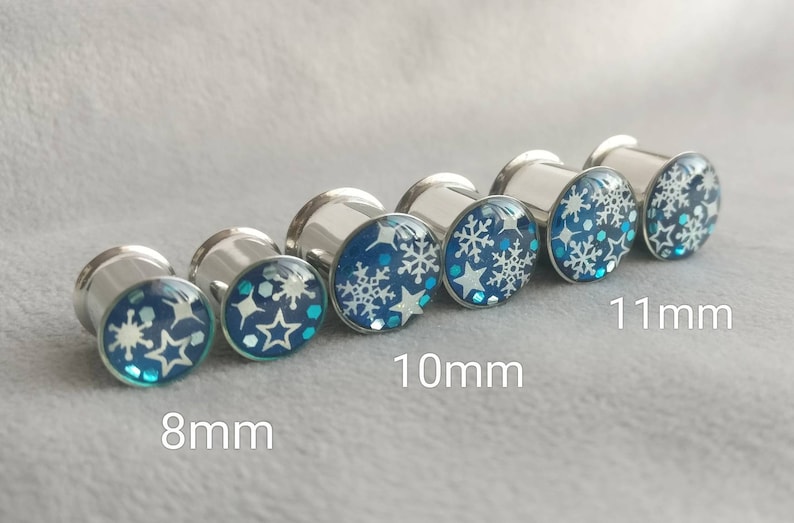 May include: Set of six silver ear gauges with blue and white snowflake designs. The gauges are 8mm, 10mm, and 11mm in size.