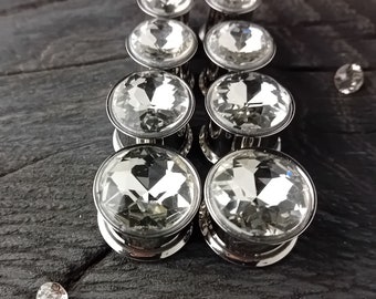 Puede incluir: Un conjunto de ocho dilatadores de oído de cristal transparente con engastes de metal plateado. Los dilatadores están dispuestos en dos filas de cuatro, con los dilatadores más grandes en la fila de atrás.
