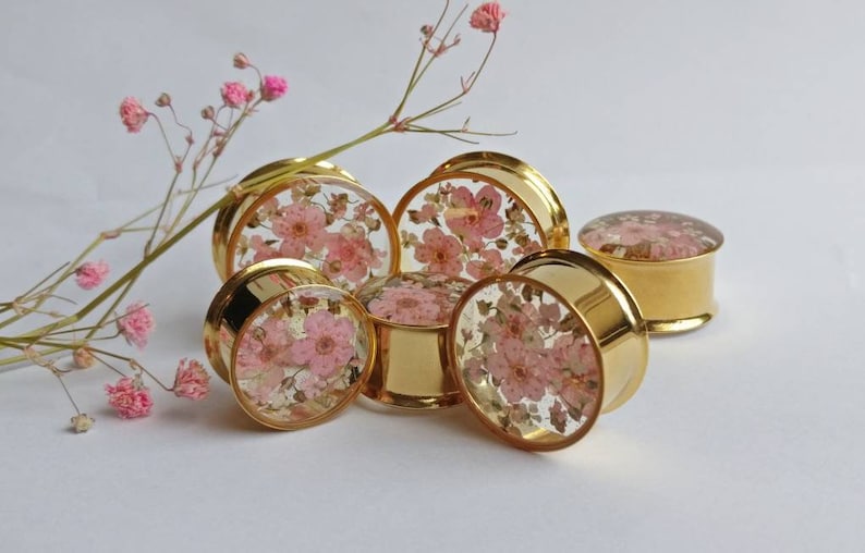 Puede incluir: Un conjunto de expansores de oreja dorados con tapas de resina transparente, cada uno con delicadas flores rosas incrustadas. Los expansores, de varios tama&ntilde;os, est&aacute;n dispuestos sobre una superficie blanca, con una ramita de flores rosas en el fondo.