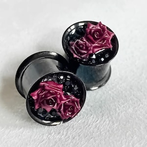 May include: Two black ear gauges featuring a dark red rose design. The roses are set within black glitter. These metal gauges have a flared shape. Suitable for stretched ear piercings, these are a unique piece of jewellery.