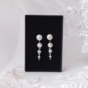 May include: A pair of silver dangle earrings with white pearl accents. The earrings feature a large pearl at the top, followed by three smaller pearls.