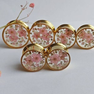 Puede incluir: Tapones para los o&iacute;dos dorados con caras de resina transparente, cada uno incrustado con delicadas flores rosas. Los tapones est&aacute;n dispuestos en grupo, mostrando su dise&ntilde;o floral. Una ramita de flores rosas est&aacute; en el fondo.