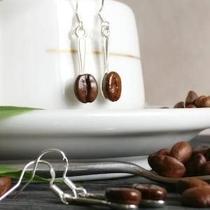 Könnte beinhalten: Silberne Ohrringe mit einem Kaffeebohnen-Anhänger. Die Ohrringe hängen an einer weißen Tasse und Untertasse. Auf einer Holzoberfläche liegen Kaffeebohnen verstreut.