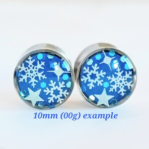 May include: Pair of 10mm (00g) stainless steel ear gauges with blue and white snowflake and star designs. 10mm (00g) example.