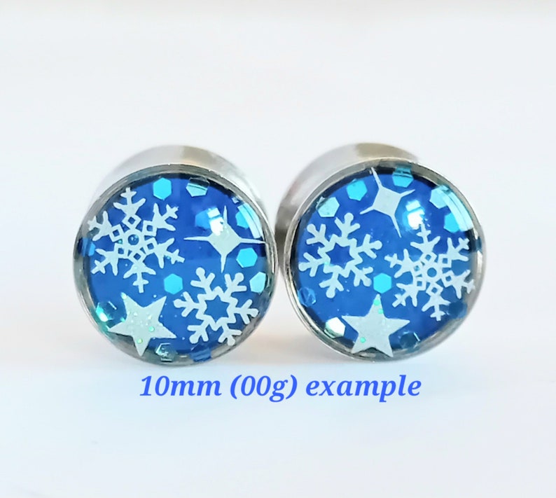 May include: Pair of 10mm (00g) stainless steel ear gauges with blue and white snowflake and star designs. 10mm (00g) example.