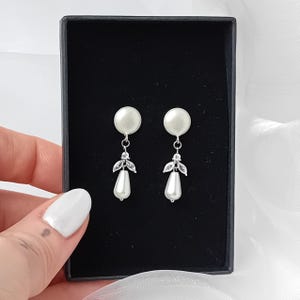 May include: A pair of silver dangle earrings with a white pearl drop and crystal accents. The earrings are displayed in a black box.