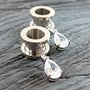 Può includere: Due dilatatori per orecchie argentati con gemme chiare a forma di goccia pendenti. I dilatatori sono cilindrici con un bordo svasato. Le gemme sono sfaccettate e tenute da un'incastonatura argentata, sospese da una delicata catena.