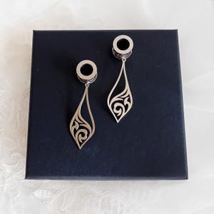 Può includere: Un paio di dilatatori per orecchie color argento con un design traforato a forma di goccia. I dilatatori sono esposti su una scatola blu scuro, con uno sfondo bianco. I dilatatori hanno una parte superiore circolare e una parte inferiore decorativa pendente.