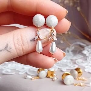 Puede incluir: Un par de pendientes de perla blanca con detalles dorados y adornos de cristal transparente. Los pendientes tienen una pequeña bola de perla blanca en la parte superior y una perla más grande en forma de pera que cuelga debajo.