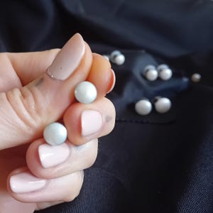 Può includere: Primo piano di una mano che tiene due orecchini rotondi e perlacei. Altri orecchini sono sparsi su un tessuto scuro. Gli orecchini hanno una lucentezza sottile, forse fatti di perle o un materiale simile.