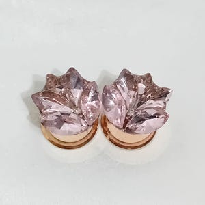May include: Two pink crystal flower-shaped ear plugs with gold-colored bases. The plugs have multiple facets that catch the light, creating a sparkling effect. The design is elegant and suitable for various occasions.