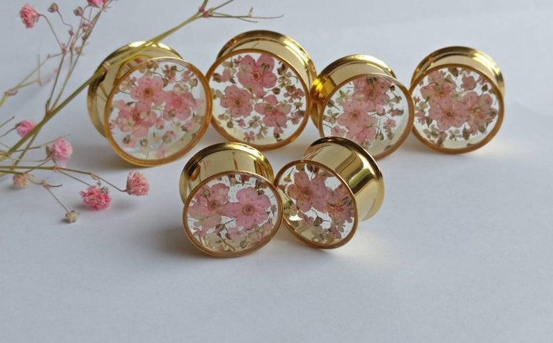 Puede incluir: Tapones para los o&iacute;dos dorados con un centro transparente lleno de flores rosas. Los tapones est&aacute;n dispuestos sobre una superficie blanca, con una ramita de flores rosas en el fondo. Un accesorio &uacute;nico.