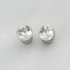 Może przedstawiać: Para przezroczystych, fasetowanych kryształowych kolczyków z podstawą w kolorze srebrnym. Kolczyki są okrągłe i mają błyszczący, odblaskowy wygląd. Tło jest jednolite, jasnoszare.