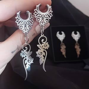 Può includere: Orecchini pendenti color argento con intricati disegni floreali e a volute. Gli orecchini presentano una parte superiore decorativa e una sezione inferiore lunga e fluente con un motivo floreale. Un secondo paio di orecchini è visibile in una scatola nera.