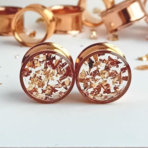 May include: A pair of rose gold-colored ear gauges with clear centers filled with gold leaf flakes. Additional gauges are visible in the background, along with scattered gold leaf pieces. The gauges have a polished, metallic finish.