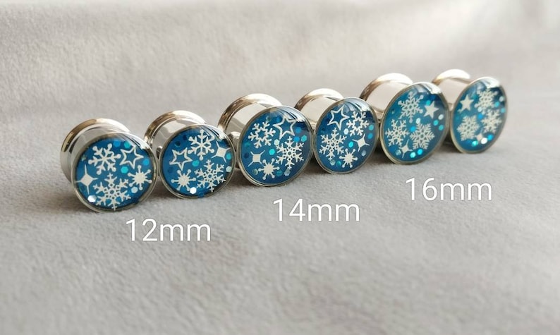 May include: Set of five silver ear gauges with blue and white snowflake designs. The gauges are 12mm, 14mm, 16mm in size.