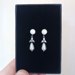 May include: A pair of elegant pearl earrings. Each earring features a round pearl stud, a decorative silver element with leaf-like details and small crystals, and a teardrop-shaped pearl drop. The earrings are displayed against a black background.