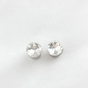 Puede incluir: Dos pendientes de botón redondos de cristal transparente con un engaste de metal plateado. Los pendientes están facetados, reflejando la luz, sobre un fondo blanco. Una joya clásica.