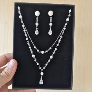 May include: A silver-toned jewellery set consisting of a double-strand necklace and matching drop earrings. The necklace features white pearls and clear faceted crystals on a delicate chain, with a central pear-shaped crystal pendant drop.