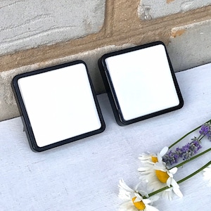 以下が含まれることがあります： 白のセンターと黒の縁取りが付いた正方形のコースターが2つ。白い表面に置かれ、ラベンダーとデイジーの小枝が前景にあります。背景はレンガの壁です。