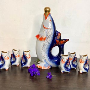 Puede incluir: Un juego de vasos de chupito de porcelana azul y blanca con forma de pez y una jarra a juego. La jarra es más alta que los vasos de chupito y tiene una tapa dorada. El juego está decorado con detalles en naranja y rojo.