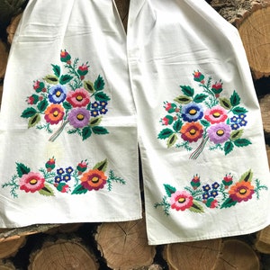 Può includere: Due pannelli di tessuto bianco con ricami floreali colorati. Il ricamo presenta bouquet di fiori nelle tonalità di rosso, arancione, blu, viola e verde. I pannelli sono esposti su uno sfondo di tronchi accatastati.