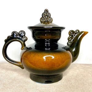 Könnte beinhalten: Eine Keramik-Teekanne mit dunkelbrauner und bernsteinfarbener Glasur. Die Teekanne hat einen dekorativen Griff, Ausguss und Deckel mit aufwendigen Details. Die Teekanne ist im Vintage-Stil gehalten und steht auf einem kleinen Sockel.