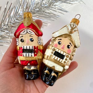 May include: Two Christmas ornaments shaped like nutcrackers. One is red and gold, the other is white and gold. Both have detailed faces, black mouths, and black boots. The ornaments are held in a hand, with a silver tinsel garland in the background.