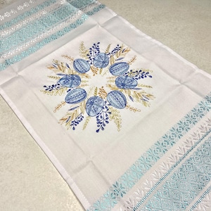 May include: White Easter table runner with blue and gold embroidered egg and floral design. The edges feature a turquoise and white woven pattern. A decorative piece for the Easter holiday.
