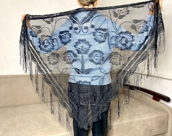 May include: A black lace shawl with a floral pattern and fringe. The shawl is held up, displaying the intricate design and delicate texture. The floral pattern is visible against a light blue background.