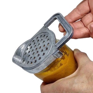 May include: A silver metal jar opener with a handle and a grater on the top. The opener is being used to open a jar of orange-colored food.