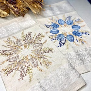 May include: Two embroidered kitchen towels with a white background and a border of small white diamonds. One towel features a wreath of blue embroidered eggs and the other features a wreath of brown embroidered eggs.