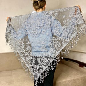 May include: A gray lace shawl with a floral pattern and fringe trim. The shawl is held up, showcasing its intricate design and delicate texture. The shawl is a triangle shape and appears to be made of a lightweight material.