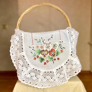 May include: A wicker basket with a high handle, lined with white fabric. The fabric is embroidered with colourful floral and Easter egg motifs, and trimmed with a delicate, scalloped lace. The basket is likely for Easter.