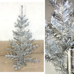 May include: A silver artificial Christmas tree with metallic tinsel needles and a central pole. The tree features tiered branches that extend outward from a central stem, creating a conical shape. The needles have a reflective, shiny finish.
