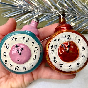 May include: Two vintage Christmas ornaments shaped like clocks. One is teal and pink, the other is red and gold. Each clock face has black numerals and hands pointing to the time. A festive holiday decoration.