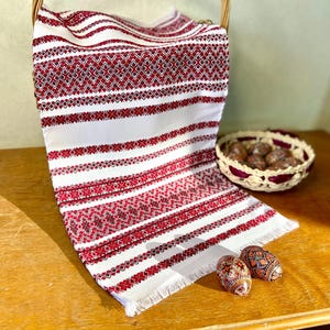 May include: White and red embroidered textile with a geometric pattern, draped over a basket. Two decorated Easter eggs are on a wooden surface. A small woven basket with brown objects is also present.