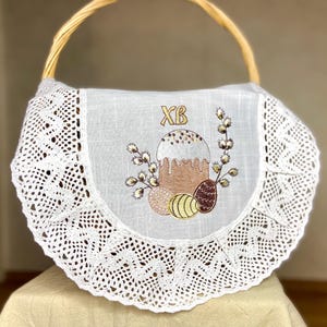 May include: A wicker basket with a white embroidered cover. The cover is decorated with an Easter cake, eggs, and pussy willow branches. The cover has a lace trim. The letters "XB" are embroidered in gold.
