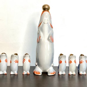 May include: A set of seven porcelain fish-shaped liquor bottles. The largest bottle is in the center and has a gold top. The smaller bottles are arranged in a row in front of the larger bottle. The bottles are decorated with orange, blue, and white paint.