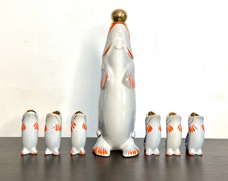May include: A set of seven porcelain fish-shaped liquor bottles. The largest bottle is in the center and has a gold top. The smaller bottles are arranged in a row in front of the larger bottle. The bottles are decorated with orange, blue, and white paint.