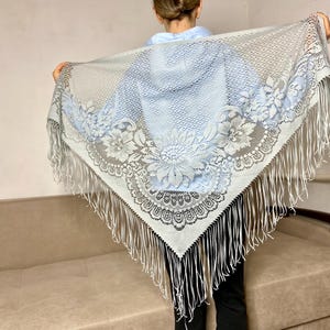 Puede incluir: Un chal triangular con un patrón de encaje floral en azul claro y blanco con un centro de malla. Los bordes están adornados con largos y finos flecos blancos que cuelgan de todo el perímetro de la tela, creando un borde decorativo.