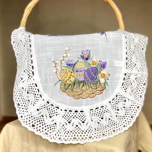 May include: A white Easter basket with a woven handle and an embroidered panel. The panel shows colourful Easter eggs, purple flowers, and a woven basket design. The basket is trimmed with delicate lace.