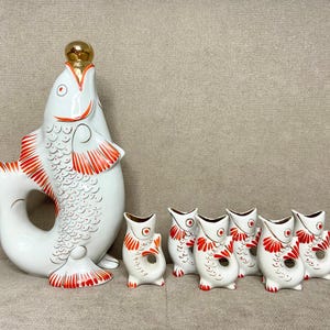 May include: A set of seven white porcelain fish-shaped decanters with red and gold accents. The largest decanter is a large fish with a gold top and the smaller decanters are smaller fish with red and gold accents.