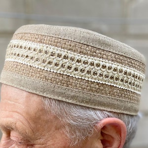 Puede incluir: Un sombrero tipo pastillero de color tostado con la parte superior plana, hecho de una tela tejida gruesa. El sombrero presenta una banda horizontal ancha de encaje calado intrincado de color crema que rodea la corona, creando contraste.