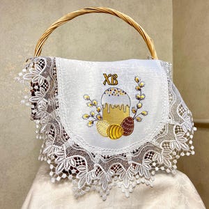 May include: A wicker basket with a curved handle. The basket is lined with a white cloth featuring an embroidered design of a cake, eggs, and the letters "XB". The cloth is trimmed with white lace.